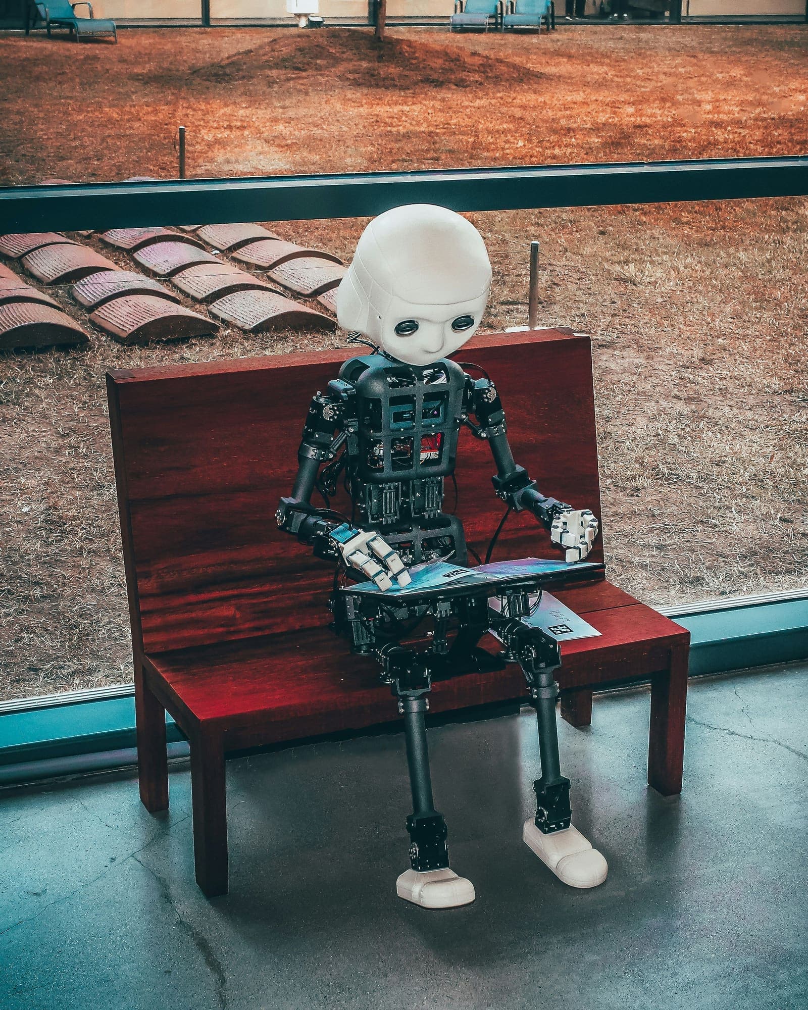 A small humanoid robot sitting on a bench, reading a tablet.