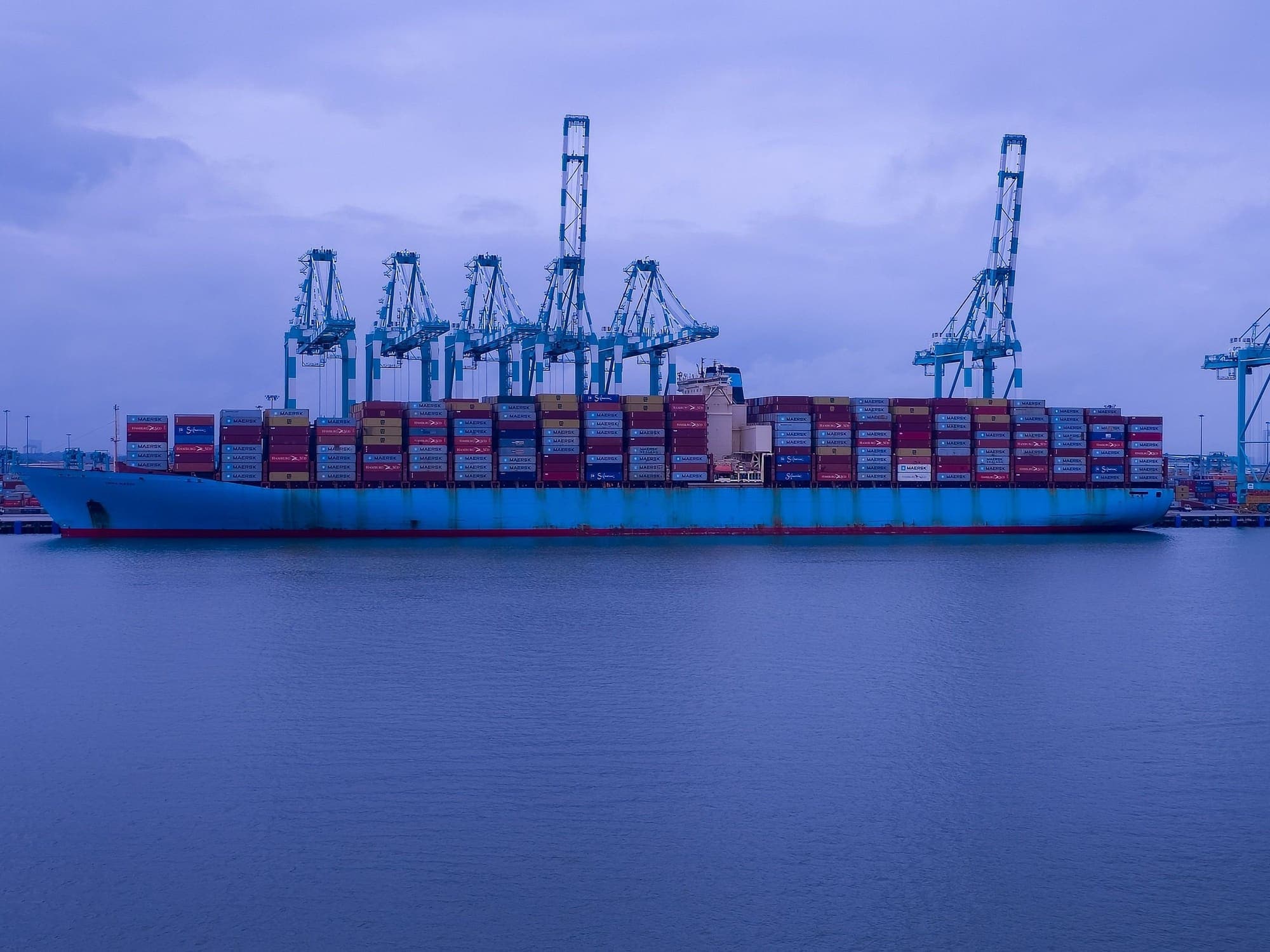 A container ship stacked with containers under port cranes at dusk.