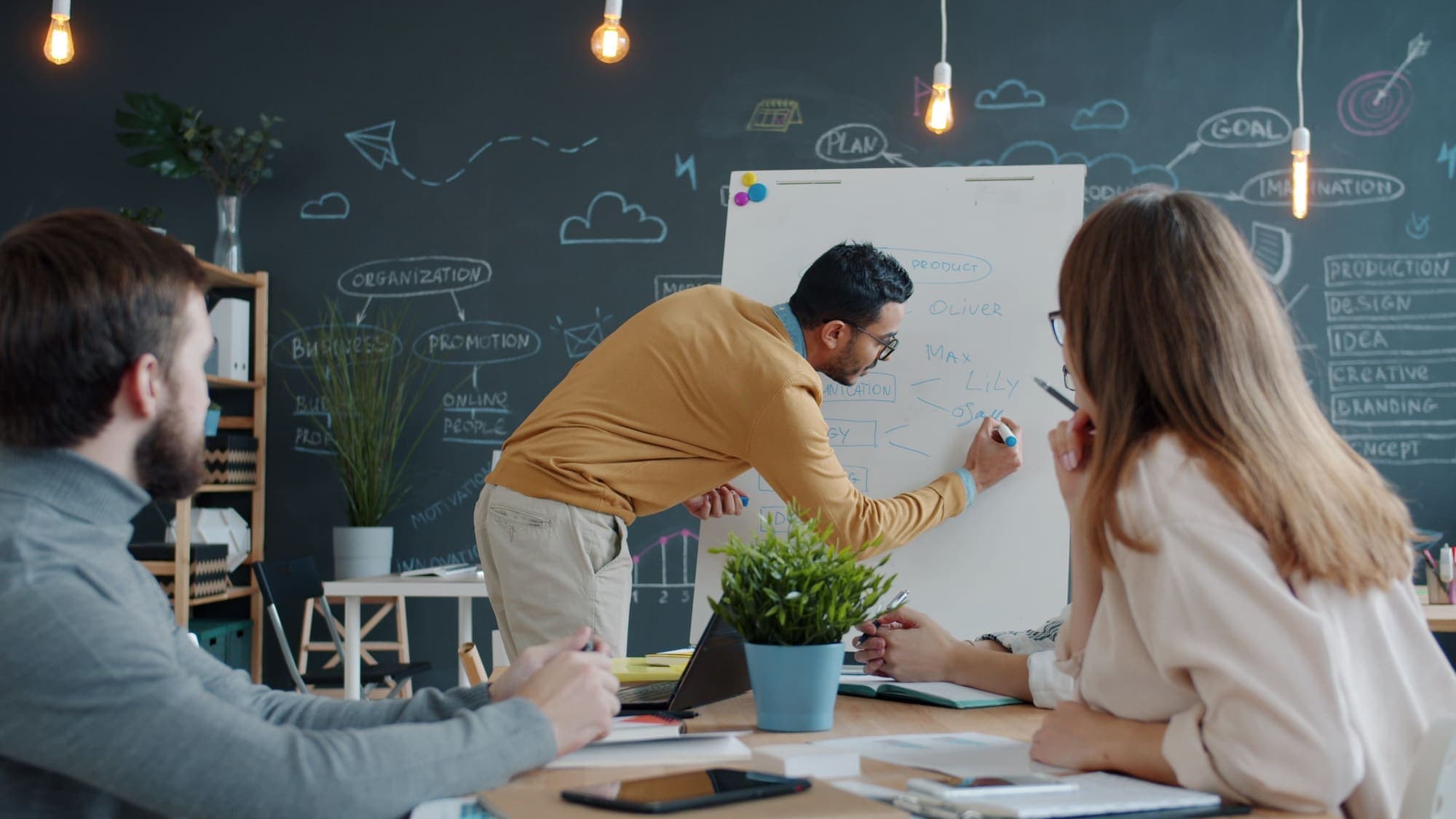 Three colleagues at a table while one writes a plan on a whiteboard.