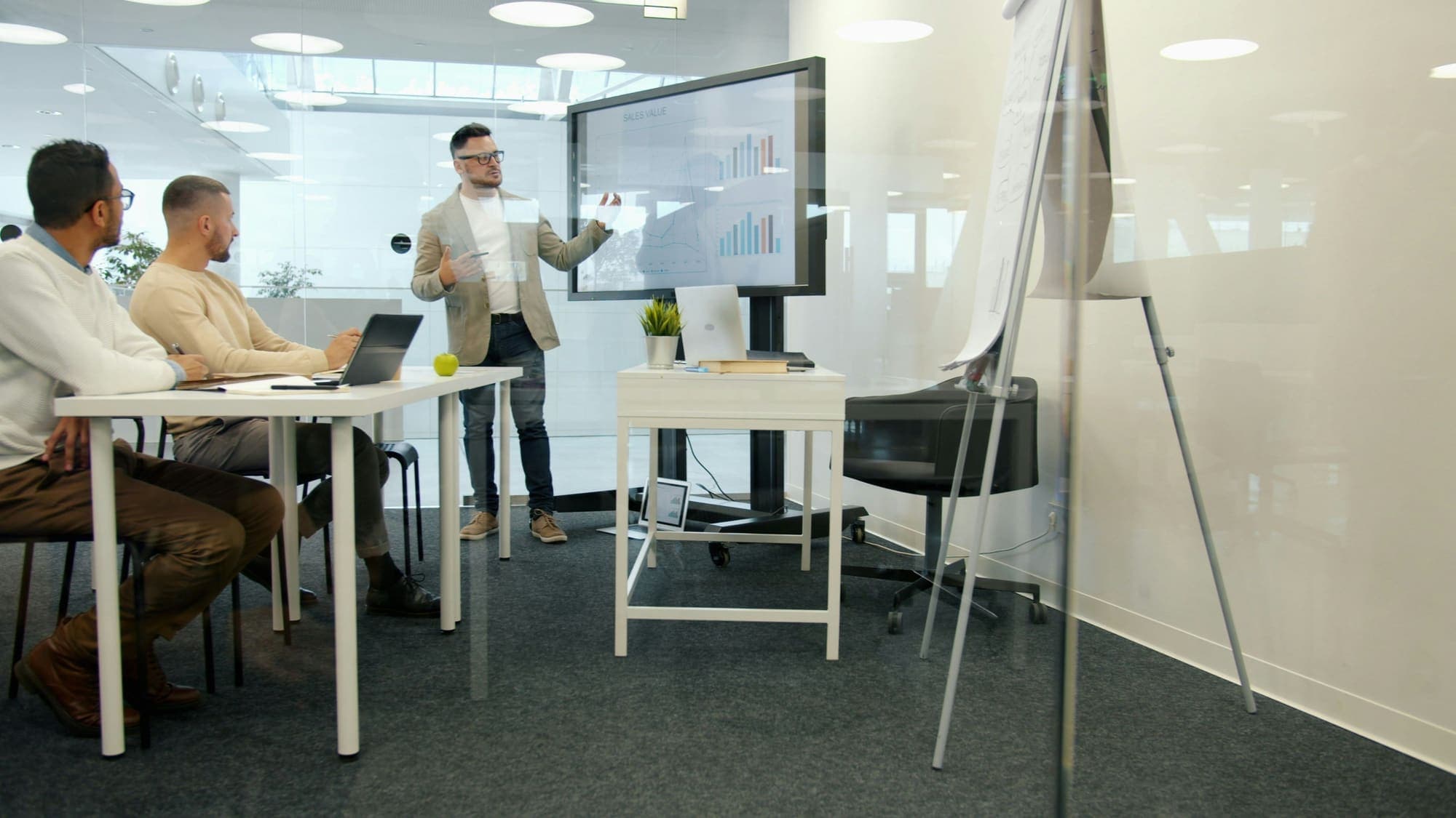 A person presents charts on a wall screen to two colleagues at a table.